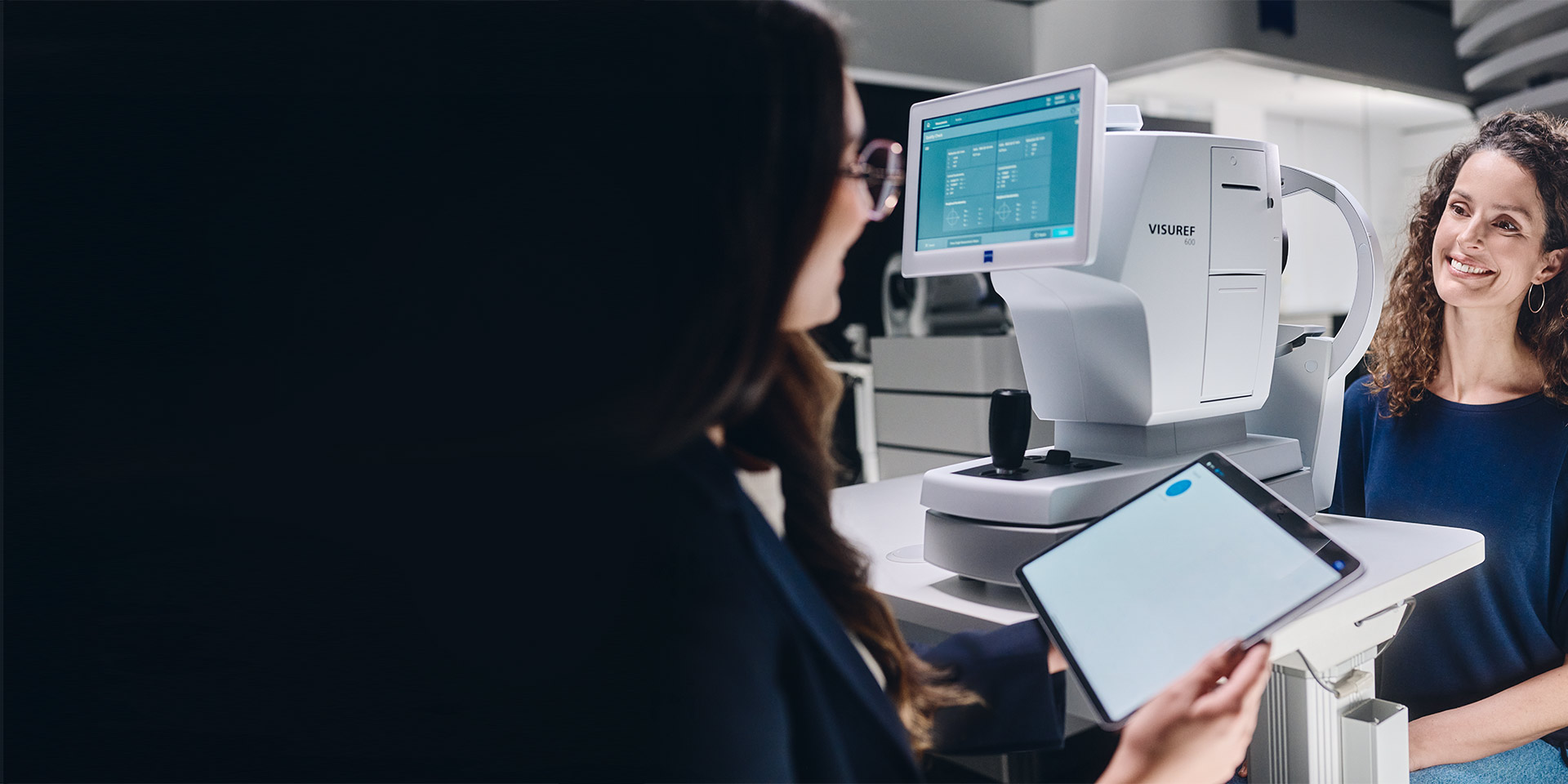 En kvindelig optiker ser på sin tablet og sidder foran ZEISS VISUREF 600 under en samtale med sin kunde.