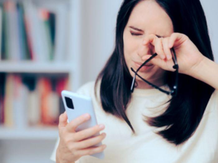 Et billede af en kvinde i 30-erne, der kigger på sin telefon, mens hun gnider sine øjne.