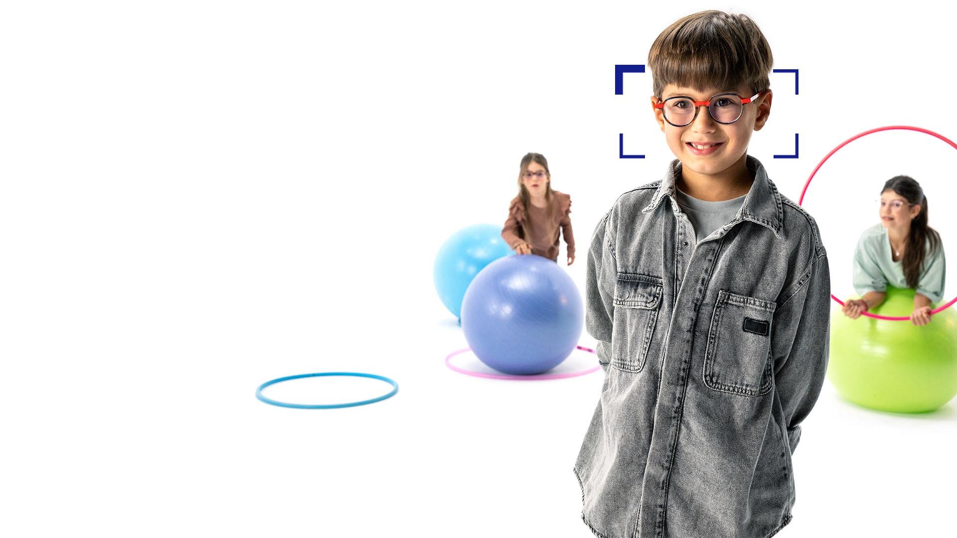 Dreng med brunt hår og runde briller med ZEISS MyoCare-brilleglas står i forgrunden og smiler til kameraet. I baggrunden ses to piger, der bærer briller med ZEISS MyoCare-brilleglas og leger med hoola-hoopringe og hoppebolde.