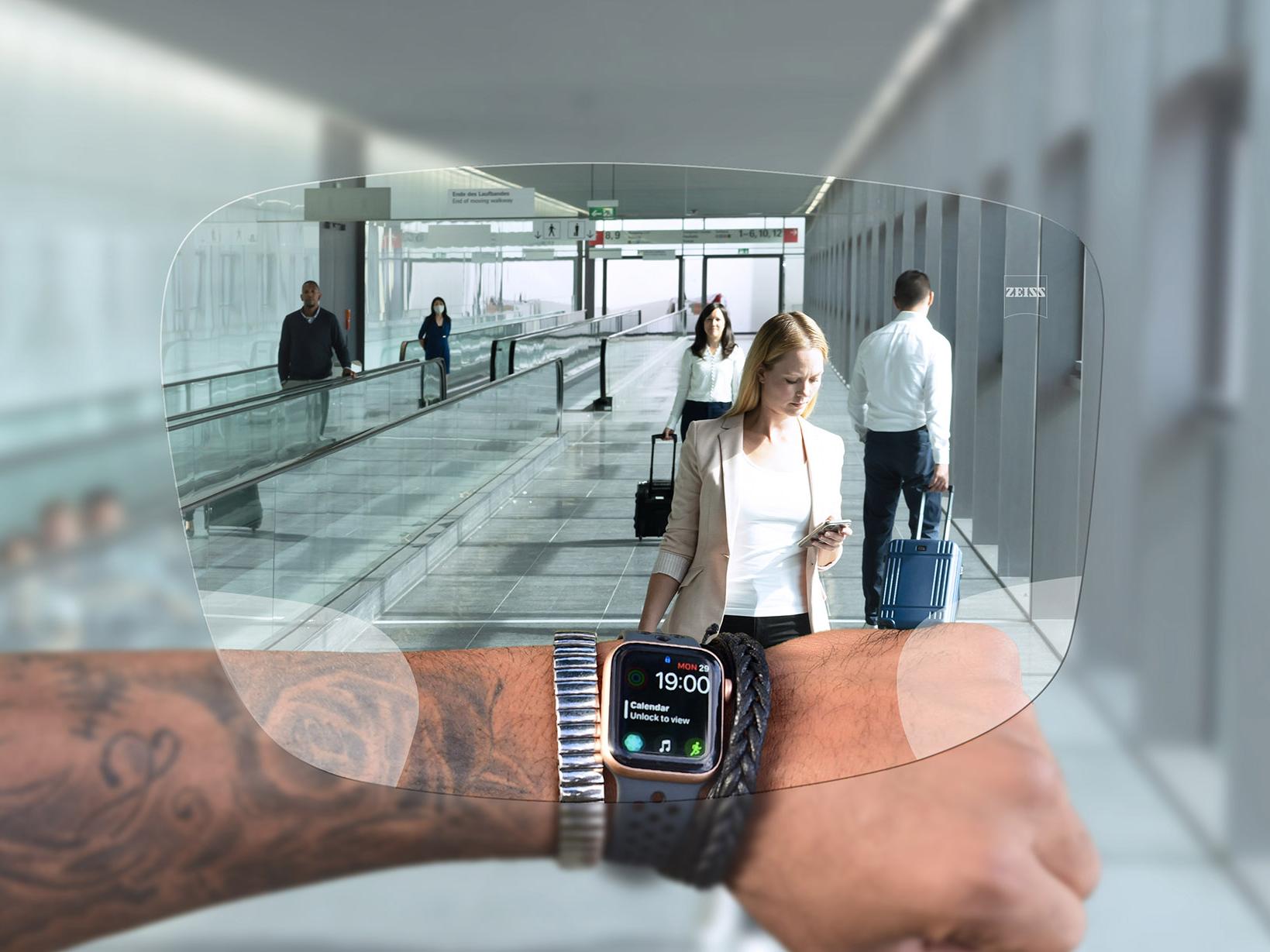 En person ser på sit ur igennem ZEISS SmartLife Digital brilleglas. I baggrunden ses en typisk situation i en lufthavn med mennesker på rulletrapper, der medbringer kufferter. 
