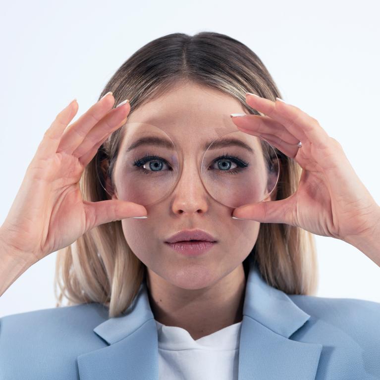 En ung blond kvinde holder brilleglas op foran øjnene for at vise det gode udseende uden nogen morsom øje-effekt, i kraft af ZEISS ClearView enkeltstyrkeglas.