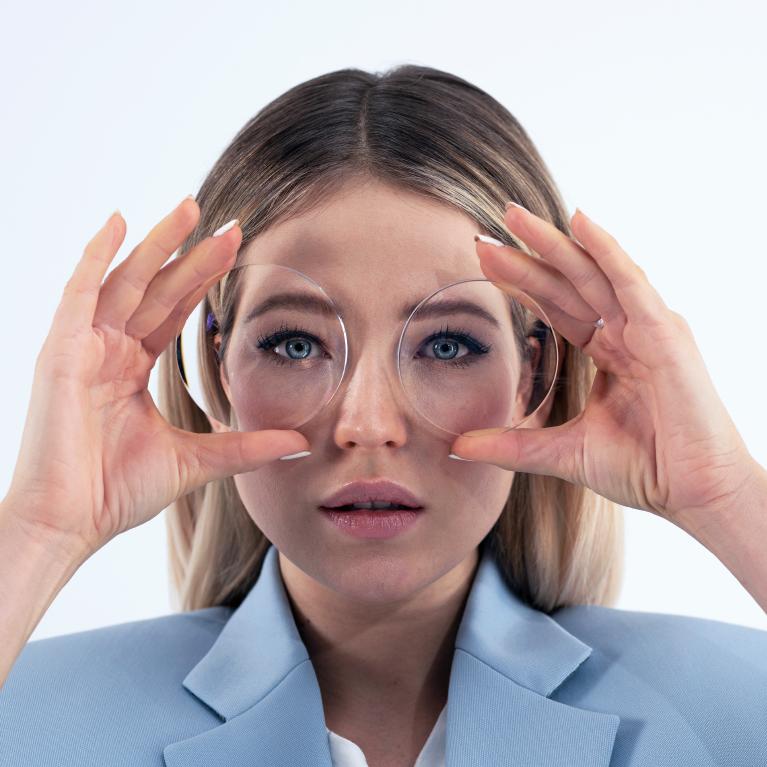 En ung blond kvinde holder brilleglas op foran øjnene for at vise miniature-øje-effekten af tykke minusglas.