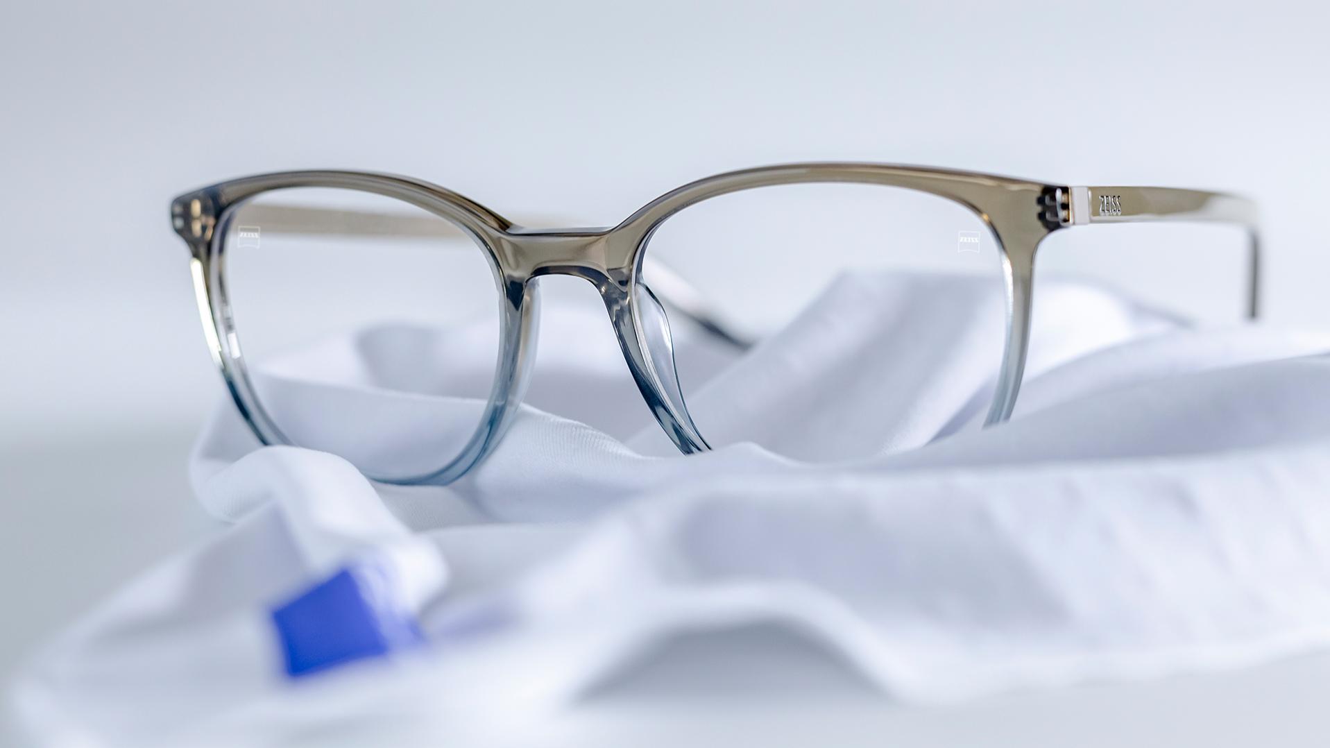 Et par briller med grå-blåt stel og ZEISS glas med DuraVision®-coating ligger på en hvid mikrofiberklud.