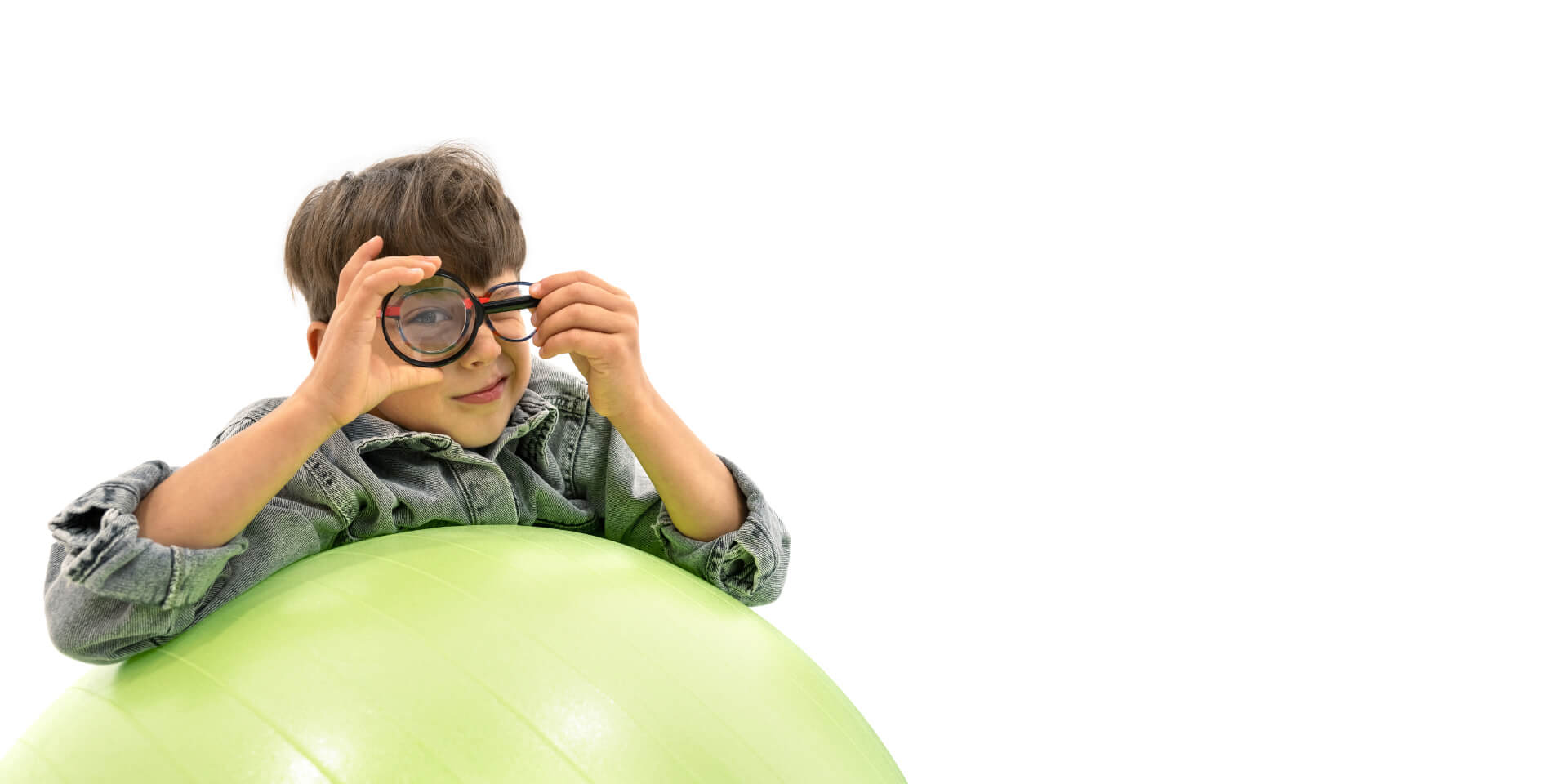 En dreng der har ZEISS briller på til kontrol af myopi, og som læner sig op af en hoppebold og holder en lup for øjet.