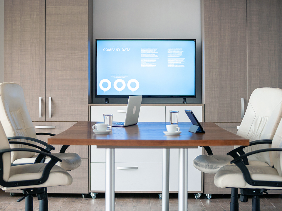 Udsigt fra skrivebordet på et chefkontor. En monitor er monteret på en væg et par meter væk. Der er et lille mødebord med kontorstole og bærbare på bordet.