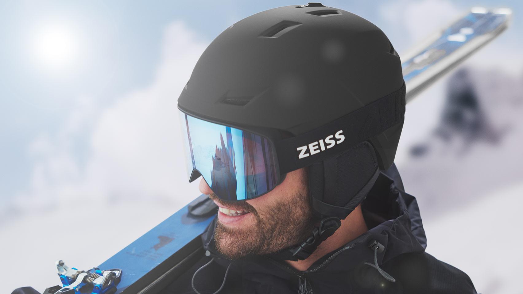 En mand har en sort hjelm på med blåspejlfarvede ZEISS skibriller og har skiene over højre skulder.