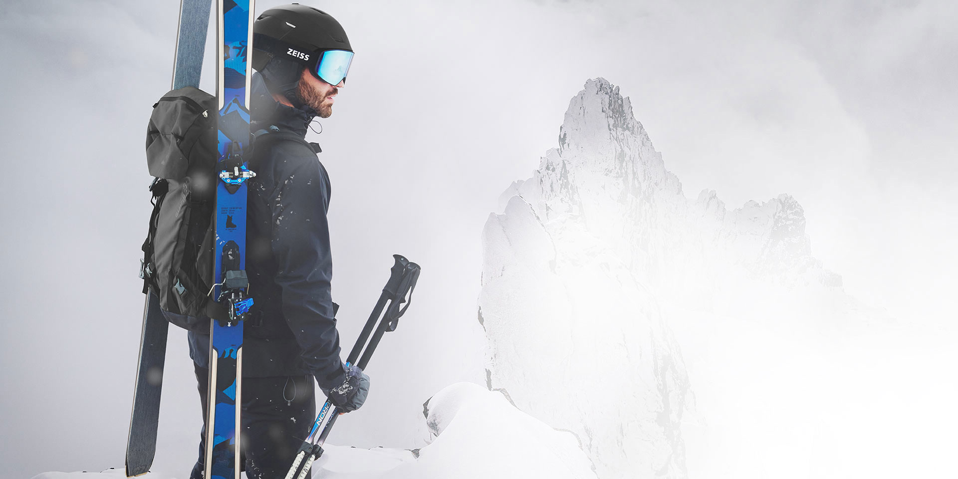 En mand har en sort hjelm på med blåspejlfarvede skibriller fra ZEISS, har ski og en rygsæk på ryggen og holder skistavene i højre hånd. Han står på toppen af et bjerg.