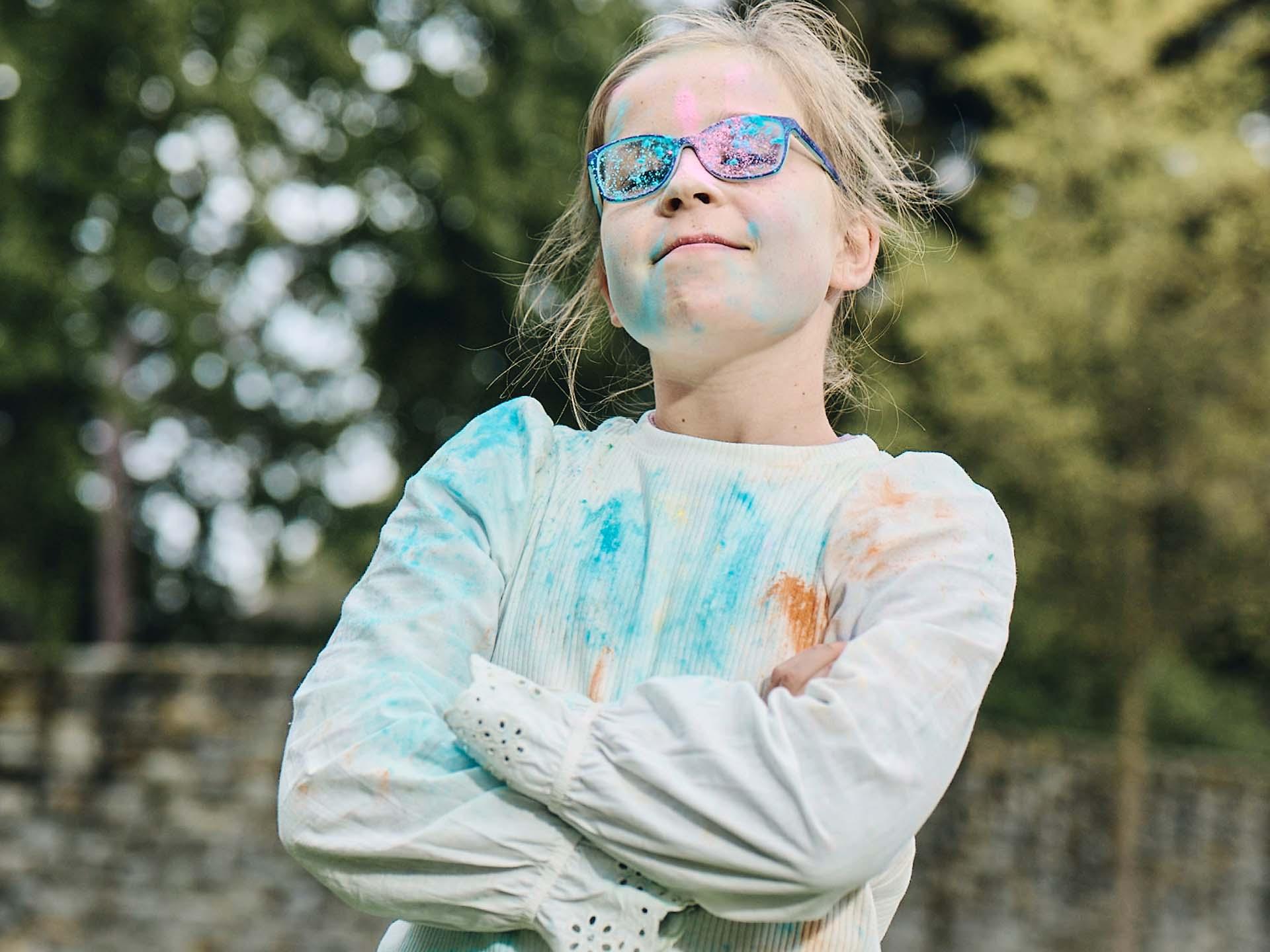 En pige med armene over kors og snavsede briller af at lege med farvet pulver ser kæk ud med et selv-tilfredst grin.