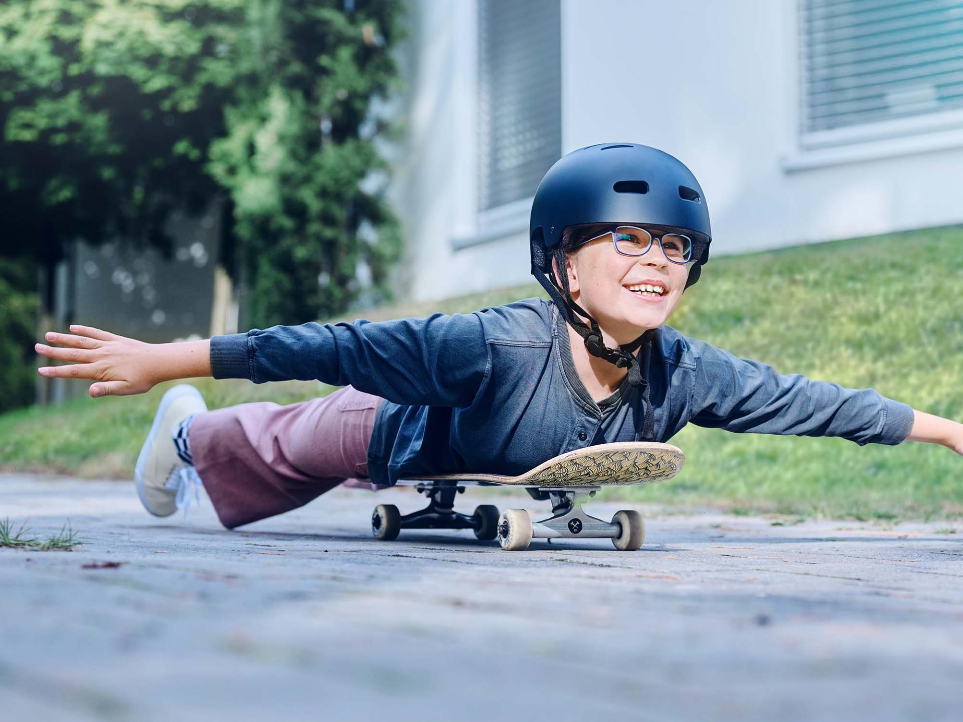 En pige med hjelm på og briller kører ned ad vejen liggende på et skateboard og strækker armene ud.