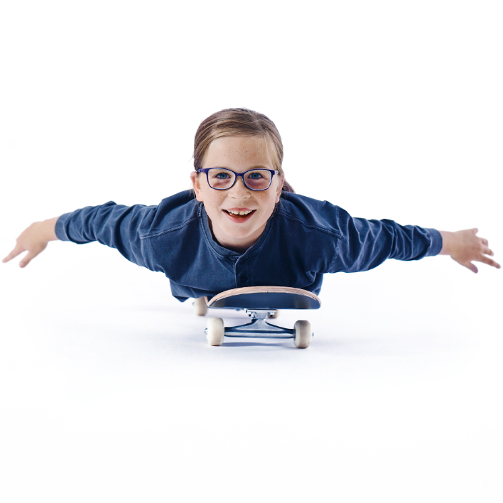 En ung pige med briller på ligger på maven på sit skateboard.